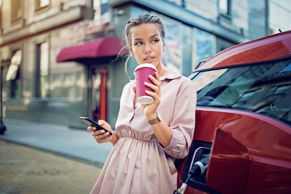 Young woman leaning against electric car with coffee in hand.